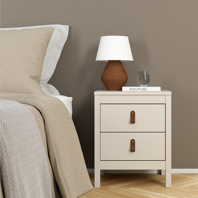 Square bedside table with two drawers, featuring leather pull handles, in a soft beige finish. A lamp and decorative