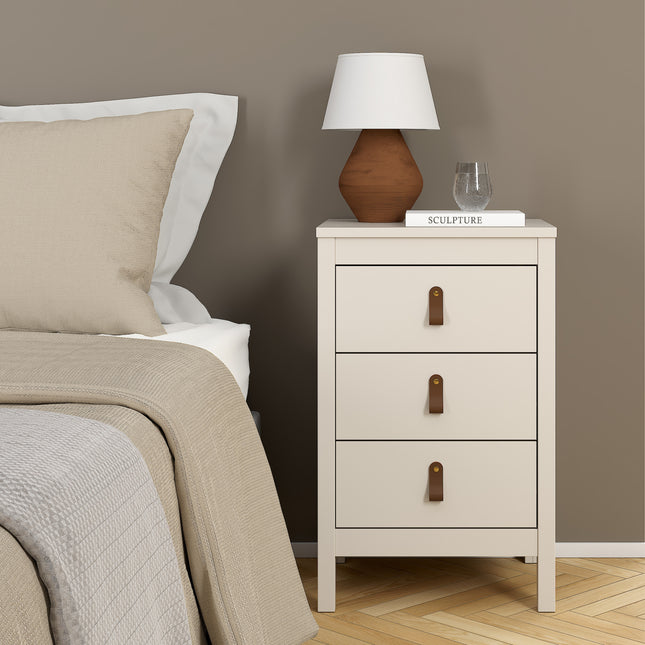 Bedside table with three drawers, light beige finish, leather pull handles, paired with a brown lamp and decorative items