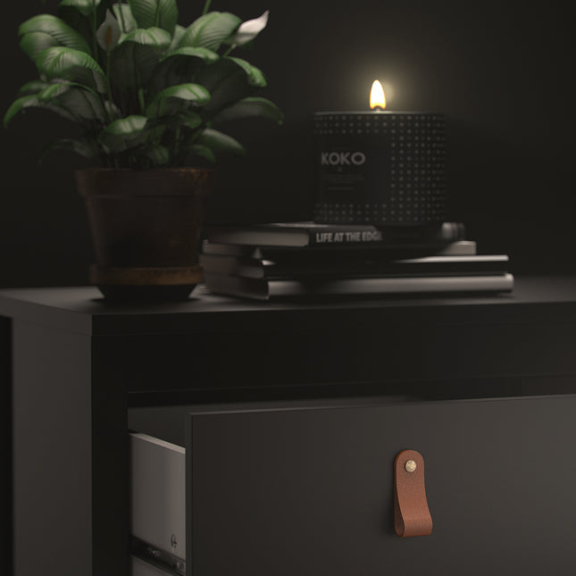Black bedside table with two drawers, featuring leather pull handles, stacked books, and a potted plant beside a lit candle