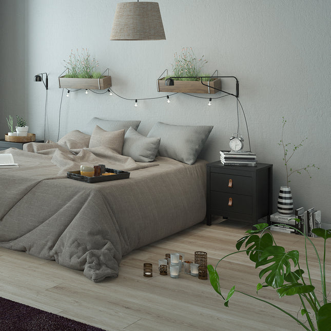Bedside table with two drawers, black finish, placed beside a bed with grey bedding and decorative plants above