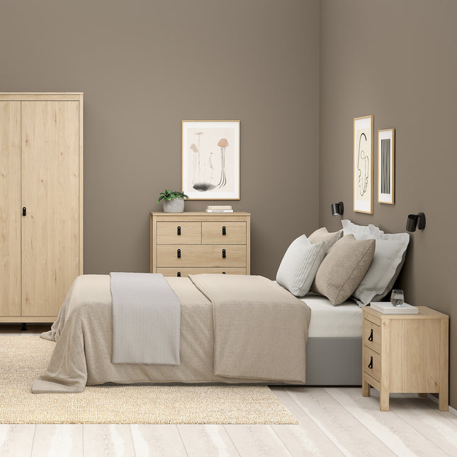 Bedside table with two drawers, light wood finish, black handles, placed beside a bed with grey bedding