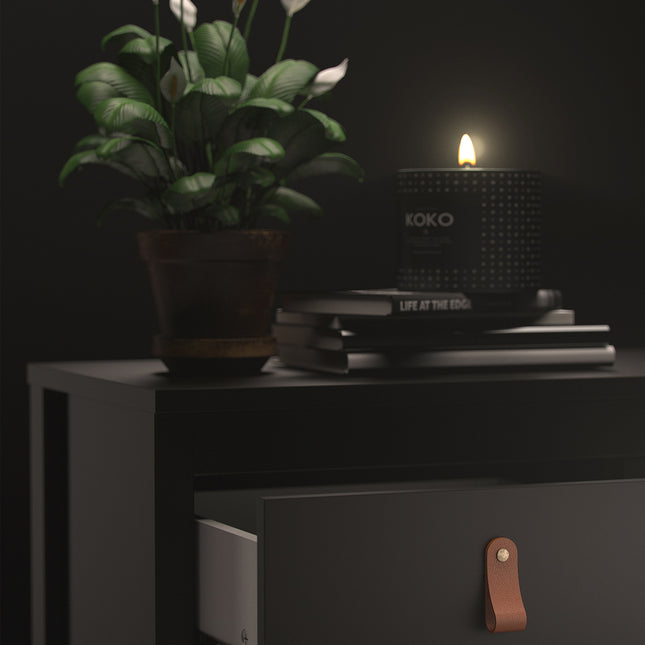 Black bedside table with a single drawer featuring a leather handle, topped with a potted plant, candle, and stacked books