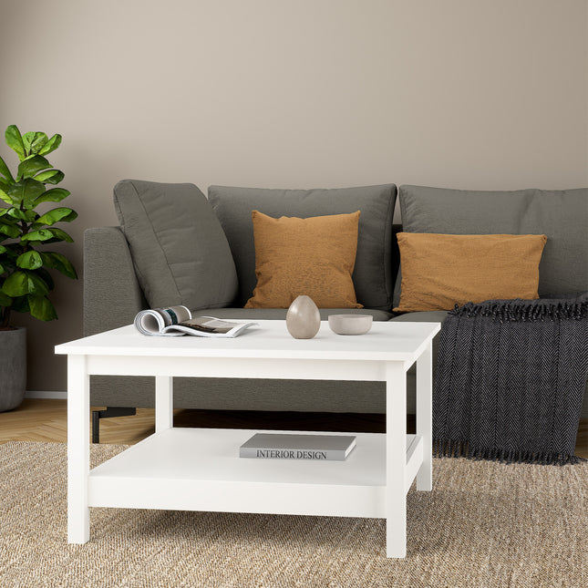 Square coffee table in white with a lower shelf, placed in front of a grey sofa adorned with orange cushions