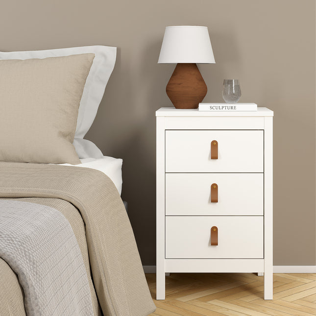 Three-drawer bedside table in white with leather pull handles, placed beside a bed with a beige quilt and lamp