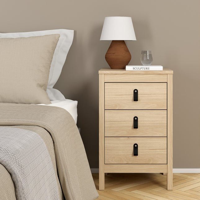 Bedside table with three drawers, light wood finish, black metal handles, beside a lamp and a decorative item on top