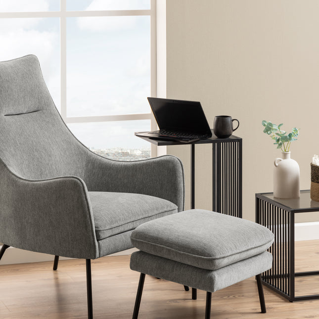 Tall side table with a sleek design, featuring vertical metal slats in black, positioned beside a grey armchair and footrest